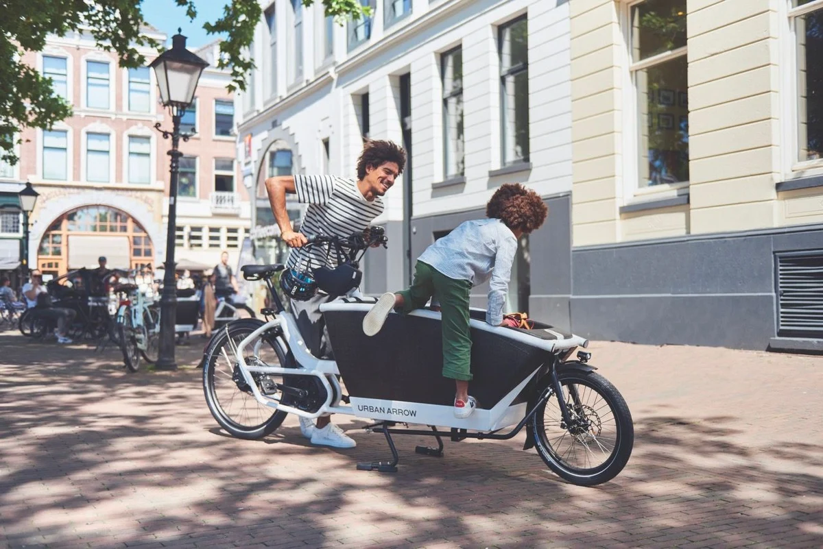 Een kind klimt in de bak van een bakfiets terwijl de vader hem vasthoudt